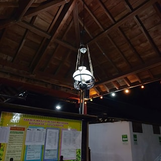 A traditional wooden ceiling with a hanging decorative lamp is featured, accompanied by a bulletin board displaying announcements and information in a public space. Colored lights are strung along near the ceiling.