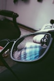 A close-up view of a pair of eyeglasses with a reflective lens. The reflection shows a window with vertical blinds, capturing a bright, blue-tinted light. The background is blurred, featuring a sneaker or shoe and a dark circular object, possibly a chair.