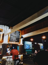 A cozy cafe interior with warm lighting. Several people can be seen sitting at tables, and a person behind the counter is preparing food. A chalkboard menu is displayed on the wall, featuring various food and drink options. In the background, there are more tables and a television screen showing a program. The overall atmosphere is casual and inviting.