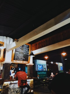 A cozy cafe interior with warm lighting. Several people can be seen sitting at tables, and a person behind the counter is preparing food. A chalkboard menu is displayed on the wall, featuring various food and drink options. In the background, there are more tables and a television screen showing a program. The overall atmosphere is casual and inviting.