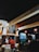 A cozy cafe interior with warm lighting. Several people can be seen sitting at tables, and a person behind the counter is preparing food. A chalkboard menu is displayed on the wall, featuring various food and drink options. In the background, there are more tables and a television screen showing a program. The overall atmosphere is casual and inviting.