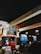A cozy cafe interior with warm lighting. Several people can be seen sitting at tables, and a person behind the counter is preparing food. A chalkboard menu is displayed on the wall, featuring various food and drink options. In the background, there are more tables and a television screen showing a program. The overall atmosphere is casual and inviting.