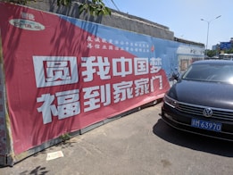 A large promotional banner with Chinese text and a red and blue color scheme is affixed to a concrete wall. A black car with a visible license plate is parked in front, partially casting a shadow on the ground.