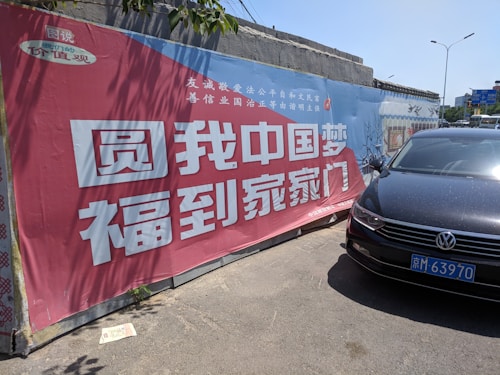 A large promotional banner with Chinese text and a red and blue color scheme is affixed to a concrete wall. A black car with a visible license plate is parked in front, partially casting a shadow on the ground.