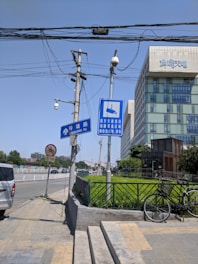 A city street scene featuring a sidewalk with traffic signs, utility poles, and surveillance cameras. On the left, a white van is parked near a 'no entry' sign. There is a blue traffic sign with Chinese characters indicating road information. A modern building with a grid-like facade is visible in the background, with greenery and a bicycle parked beside a metal fence in the foreground.