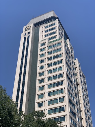 A tall, modern office building with a clock at the top, featuring numerous windows and a sleek design. The building is framed against a clear blue sky with a few trees visible at the base.