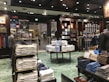 A well-organized clothing store with displays of neatly stacked shirts in various colors, mainly white, blue, and beige. Mannequins dressed in formal attire are placed around the store. Shelves are stocked with boxes and additional clothing items. Overhead lighting creates a warm atmosphere, and signs displaying prices are visible throughout the store.