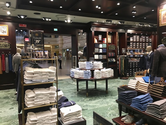 A well-organized clothing store with displays of neatly stacked shirts in various colors, mainly white, blue, and beige. Mannequins dressed in formal attire are placed around the store. Shelves are stocked with boxes and additional clothing items. Overhead lighting creates a warm atmosphere, and signs displaying prices are visible throughout the store.