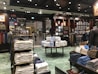 A well-organized clothing store with displays of neatly stacked shirts in various colors, mainly white, blue, and beige. Mannequins dressed in formal attire are placed around the store. Shelves are stocked with boxes and additional clothing items. Overhead lighting creates a warm atmosphere, and signs displaying prices are visible throughout the store.