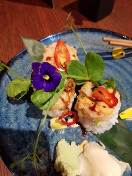 A beautifully arranged sushi plate garnished with vibrant purple flowers, green leaves, and slices of red chili pepper. The sushi pieces are topped with a creamy sauce, and there are small portions of wasabi and pickled ginger on the side. The plate is a textured blue ceramic, enhancing the presentation.