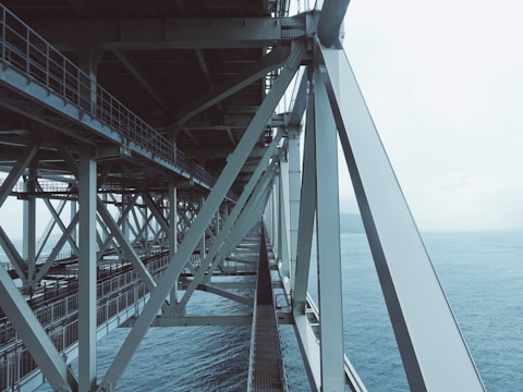A steel bridge with intricate metal support beams is seen extending over a body of water. The structure displays a network of sturdy, intersecting beams, with the ocean visible beneath and to the side. The sky above is cloudy, creating a muted, atmospheric backdrop.