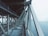 A steel bridge with intricate metal support beams is seen extending over a body of water. The structure displays a network of sturdy, intersecting beams, with the ocean visible beneath and to the side. The sky above is cloudy, creating a muted, atmospheric backdrop.