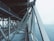 A steel bridge with intricate metal support beams is seen extending over a body of water. The structure displays a network of sturdy, intersecting beams, with the ocean visible beneath and to the side. The sky above is cloudy, creating a muted, atmospheric backdrop.