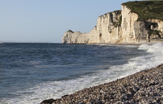 A rugged coastline features tall, white chalk cliffs with green vegetation on top. The cliffs stretch along a blue ocean, with waves gently crashing onto a stony beach.