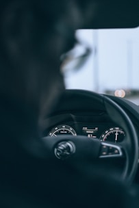 A close-up of a car dashboard, focusing on the speedometer, odometer, and fuel consumption display. The blurred foreground suggests the presence of a driver.