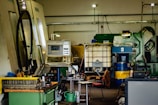 A workshop or industrial setting filled with machinery and equipment. A CNC machine with a control panel is prominent, surrounded by various tools, workbenches with scattered items, large industrial containers, and other machinery. The setting appears cluttered and functional, indicative of an active workspace.