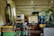 A workshop or industrial setting filled with machinery and equipment. A CNC machine with a control panel is prominent, surrounded by various tools, workbenches with scattered items, large industrial containers, and other machinery. The setting appears cluttered and functional, indicative of an active workspace.