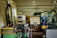 A workshop or industrial setting filled with machinery and equipment. A CNC machine with a control panel is prominent, surrounded by various tools, workbenches with scattered items, large industrial containers, and other machinery. The setting appears cluttered and functional, indicative of an active workspace.