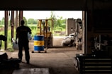 A silhouetted person stands inside a dimly lit warehouse, observing a forklift carrying a blue and yellow barrel. The open doorway reveals a bright, green outdoor scene with trees and equipment scattered around.