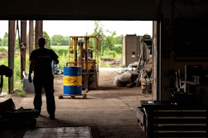 A silhouetted person stands inside a dimly lit warehouse, observing a forklift carrying a blue and yellow barrel. The open doorway reveals a bright, green outdoor scene with trees and equipment scattered around.