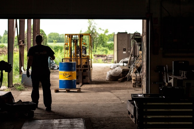 A silhouetted person stands inside a dimly lit warehouse, observing a forklift carrying a blue and yellow barrel. The open doorway reveals a bright, green outdoor scene with trees and equipment scattered around.