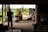 A silhouetted person stands inside a dimly lit warehouse, observing a forklift carrying a blue and yellow barrel. The open doorway reveals a bright, green outdoor scene with trees and equipment scattered around.