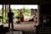 A silhouetted person stands inside a dimly lit warehouse, observing a forklift carrying a blue and yellow barrel. The open doorway reveals a bright, green outdoor scene with trees and equipment scattered around.