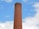 A tall, cylindrical brick chimney with metal rungs running vertically along its side, set against a bright blue sky with scattered white clouds.