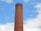 A tall, cylindrical brick chimney with metal rungs running vertically along its side, set against a bright blue sky with scattered white clouds.