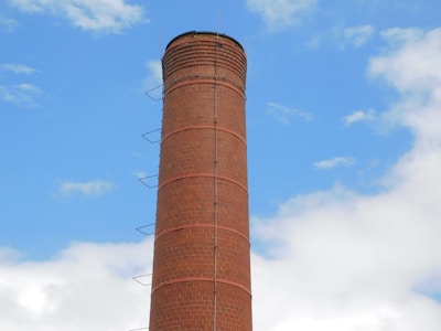 A tall, cylindrical brick chimney with metal rungs running vertically along its side, set against a bright blue sky with scattered white clouds.