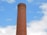 A tall, cylindrical brick chimney with metal rungs running vertically along its side, set against a bright blue sky with scattered white clouds.