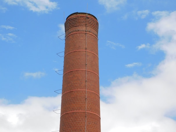 A tall, cylindrical brick chimney with metal rungs running vertically along its side, set against a bright blue sky with scattered white clouds.