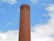 A tall, cylindrical brick chimney with metal rungs running vertically along its side, set against a bright blue sky with scattered white clouds.