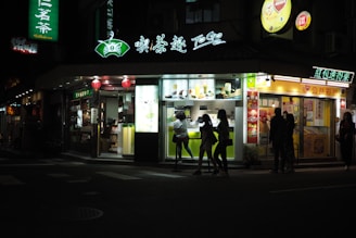 A brightly lit tea shop at night with several people standing outside. The storefront features illuminated signs with both text and images of drinks and food. Red lanterns are hung above the entrance, adding to the ambiance. A group of people can be seen silhouetted against the light from the shop, indicating a lively evening setting.