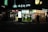 A brightly lit tea shop at night with several people standing outside. The storefront features illuminated signs with both text and images of drinks and food. Red lanterns are hung above the entrance, adding to the ambiance. A group of people can be seen silhouetted against the light from the shop, indicating a lively evening setting.