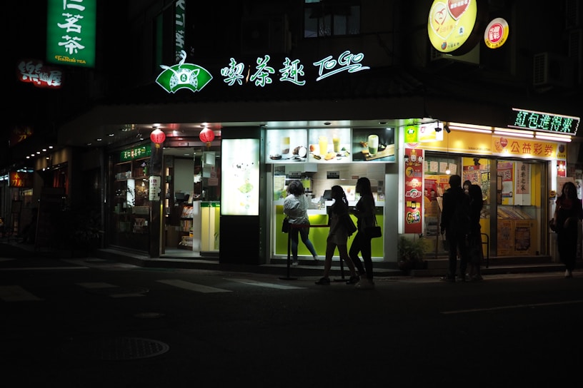 A brightly lit tea shop at night with several people standing outside. The storefront features illuminated signs with both text and images of drinks and food. Red lanterns are hung above the entrance, adding to the ambiance. A group of people can be seen silhouetted against the light from the shop, indicating a lively evening setting.