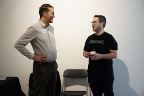 Two people are engaged in conversation in a minimalistic indoor setting. One person stands with hands on hips, while the other holds a cup, appearing engaged and smiling.