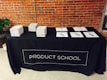 A black tablecloth with the words 'PRODUCT SCHOOL' covers a rectangular table. Stacks of books are neatly arranged on the left side of the table, while several papers with pens are laid out on the right. The backdrop features an industrial-style brick wall, and a plant can be seen to the side of the table.