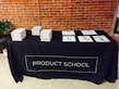 A black tablecloth with the words 'PRODUCT SCHOOL' covers a rectangular table. Stacks of books are neatly arranged on the left side of the table, while several papers with pens are laid out on the right. The backdrop features an industrial-style brick wall, and a plant can be seen to the side of the table.