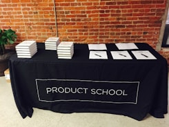 A black tablecloth with the words 'PRODUCT SCHOOL' covers a rectangular table. Stacks of books are neatly arranged on the left side of the table, while several papers with pens are laid out on the right. The backdrop features an industrial-style brick wall, and a plant can be seen to the side of the table.