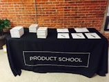 A black tablecloth with the words 'PRODUCT SCHOOL' covers a rectangular table. Stacks of books are neatly arranged on the left side of the table, while several papers with pens are laid out on the right. The backdrop features an industrial-style brick wall, and a plant can be seen to the side of the table.