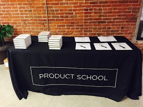 A black tablecloth with the words 'PRODUCT SCHOOL' covers a rectangular table. Stacks of books are neatly arranged on the left side of the table, while several papers with pens are laid out on the right. The backdrop features an industrial-style brick wall, and a plant can be seen to the side of the table.