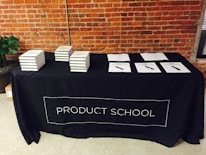 A black tablecloth with the words 'PRODUCT SCHOOL' covers a rectangular table. Stacks of books are neatly arranged on the left side of the table, while several papers with pens are laid out on the right. The backdrop features an industrial-style brick wall, and a plant can be seen to the side of the table.