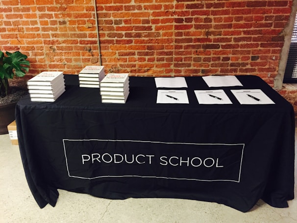 A black tablecloth with the words 'PRODUCT SCHOOL' covers a rectangular table. Stacks of books are neatly arranged on the left side of the table, while several papers with pens are laid out on the right. The backdrop features an industrial-style brick wall, and a plant can be seen to the side of the table.
