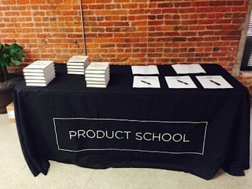 A black tablecloth with the words 'PRODUCT SCHOOL' covers a rectangular table. Stacks of books are neatly arranged on the left side of the table, while several papers with pens are laid out on the right. The backdrop features an industrial-style brick wall, and a plant can be seen to the side of the table.