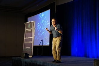 A person is standing on a stage in a dimly lit room, appearing to give a presentation. The person is animated, gesturing with both hands, and an expression of enthusiasm is evident. To the left, there's a tall banner with the words 'PRODUCT SCHOOL' and 'SILICON VALLEY'. In the background, a large screen displays a blue-toned image.