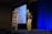 A person is standing on a stage in a dimly lit room, appearing to give a presentation. The person is animated, gesturing with both hands, and an expression of enthusiasm is evident. To the left, there's a tall banner with the words 'PRODUCT SCHOOL' and 'SILICON VALLEY'. In the background, a large screen displays a blue-toned image.