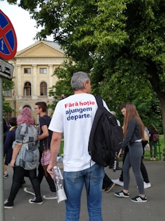 A group of people walking on a sidewalk near a historic building. The building has an ornate architectural style with columns and decorative details, partially obscured by green trees. A prominent 'no parking' traffic sign is in the foreground. One person in the center is wearing a white t-shirt with Romanian text on the back and holding some rolled papers. The scene appears to be set in a city environment with a mix of casual foot traffic.