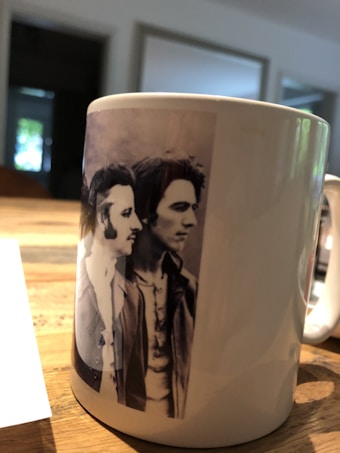 A white ceramic mug sits on a wooden table, displaying a black and white photograph of two individuals with medium length hair and sideburns. The room in the background appears to have a soft, natural lighting and blurred elements like windows and furniture.