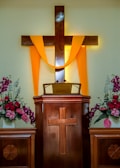 A wooden pulpit with a cross design is adorned with a bright yellow cloth draped over a larger wooden cross. Flower arrangements with pink and white blossoms are placed on each side, contributing to a serene and solemn atmosphere.